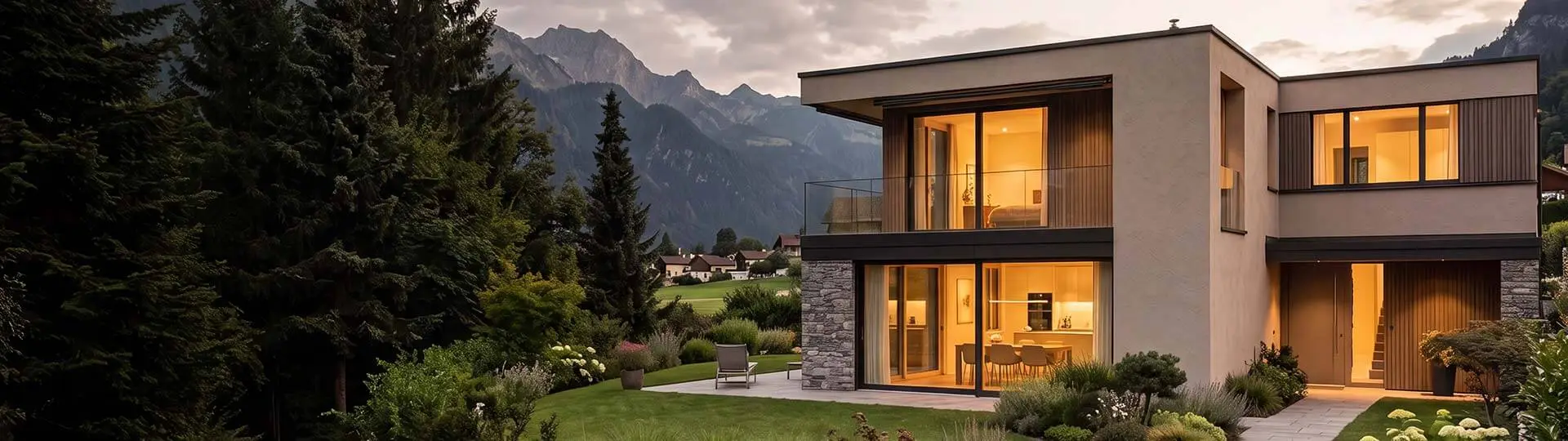 Modernes zweistöckiges Einfamilienhaus mit grossen Fenstern, Natursteinfassade und gepflegtem Garten bei Abendlicht, im Hintergrund Berge