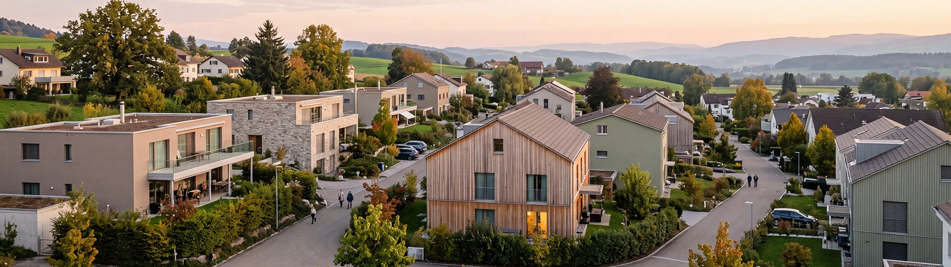 Erhöhter Blick über ein ruhiges Wohnquartier im Aargau bei Abendlicht, mit modernen Häusern mit Naturstein- und Holzfassaden, einzelnen erleuchteten Fenstern und sanften Hügeln am Horizont